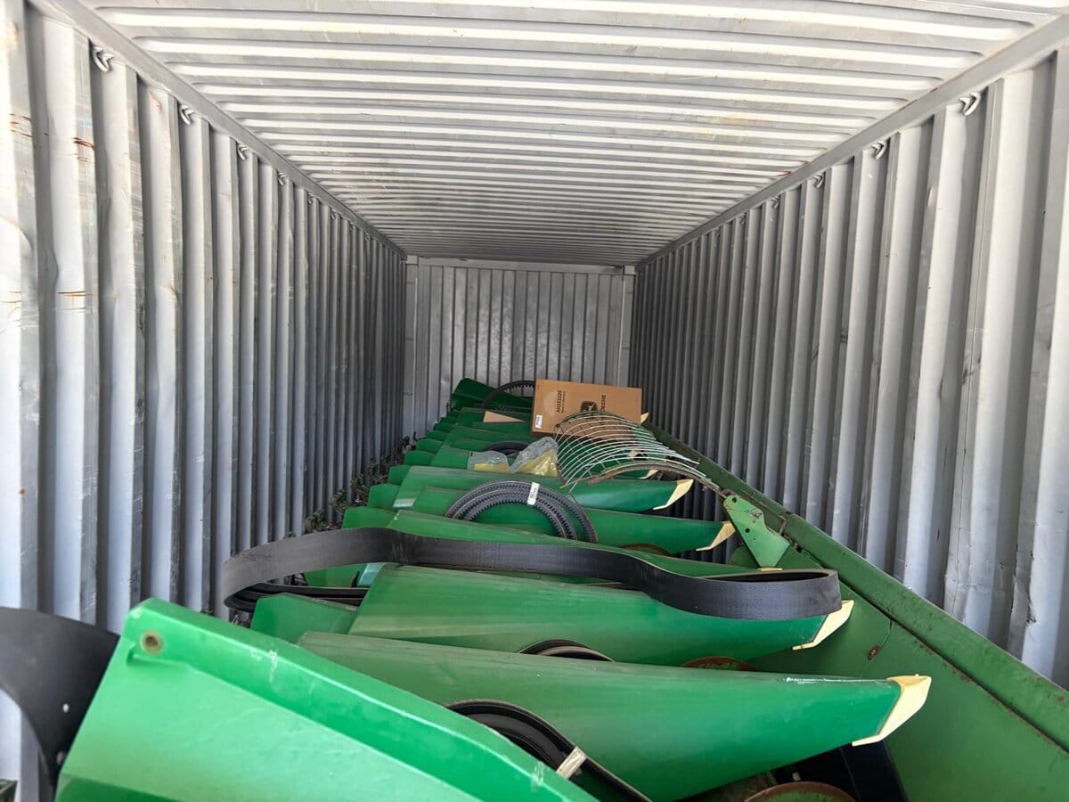 Corn header with its green row units lined up inside a shipping container, drive belts and a cardboard box at the far end